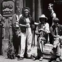 Group of men and womensit and stand next to a wooden totem pole