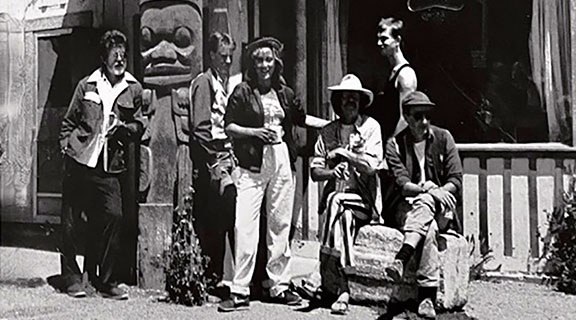 Group of men and womensit and stand next to a wooden totem pole